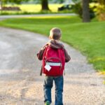 boy in brown hoodie carrying red backpack while walking on dirt road near tall trees